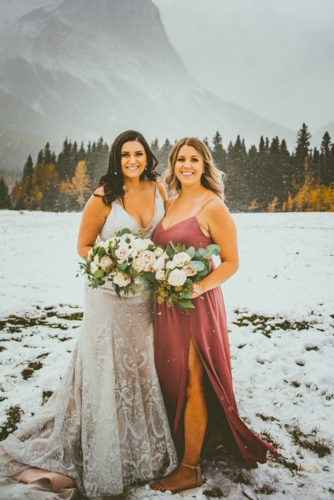Blush and Mauve Canmore Wedding - Meagan and her maid of honour standing in a snow covered field in the Rocky Mountains. The bride is wearing a champagne and ivory lace bridal gown and carrying a stunning bouquet with greenery and blush, mauve and white flowers including roses, panda anemones and ranunculus. Her maid of honour is wearing a floor-length mauve dress and carrying a simpler blush, white and mauve bouquet.