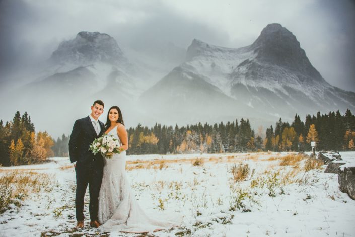 Blush and Mauve Canmore Wedding - Bride and groom standing in a snowy field in the Rocky Mountains near Canmore. Bride is carrying a blush, white and mauve bouquet.