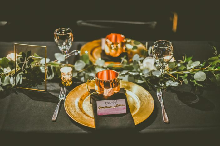Black and gold decorated reception table with a fresh eucalyptus garland. Engraved moscow mule mugs were placed on each plate.