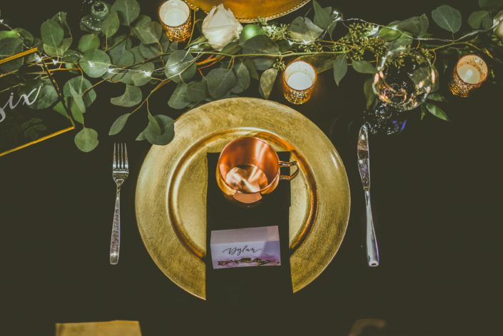 Black and gold table setting with a moscow mule mug as a wedding favour. A fresh eucalyptus greenery garland with twinkly lights ran down the centre of the table.