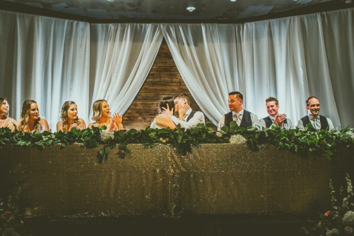 Head table decorated with a gold sparkly tablecloth and a long fresh greenery garland with fairy lights.