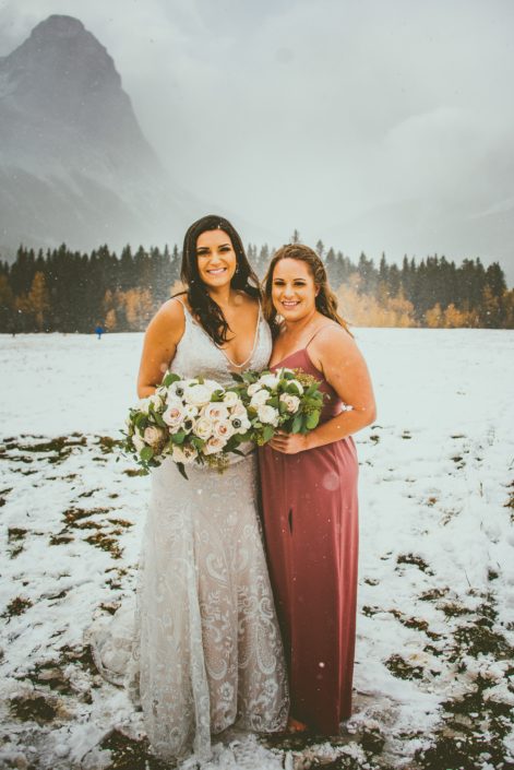 Bride and bridesmaid carrying blush bouquets standing in a snowy field in the Rocky Mountains.