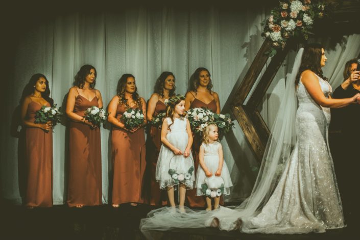Bridesmaids wearing mauve dresses and holding blush and mauve bouquets standing with flower girls wearing flower crowns and carrying floral hoops. Hexagon archway decorated with an arrangement made of blush, mauve and white flowers and greenery.
