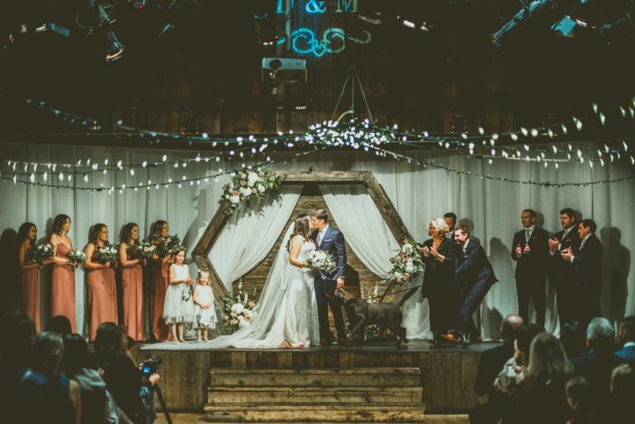 Meagan and Dwayne kiss under a wooden hexagon archway decorated with white, mauve and blush flowers with greenery at their wedding ceremony and are congratulated by their excited dog.