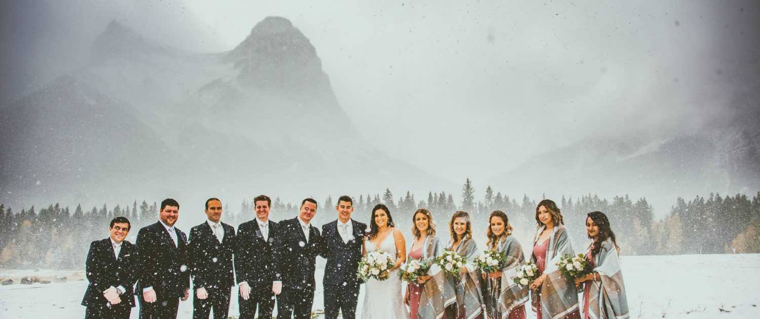 Blush and Mauve Canmore Wedding - Meagan and Dwayne's Bridal Party standing in the snowy Rocky Mountains at Canmore. The bridesmaids are wearing mauve dresses and shawls and holding mauve, blush and white bouquets with greenery.