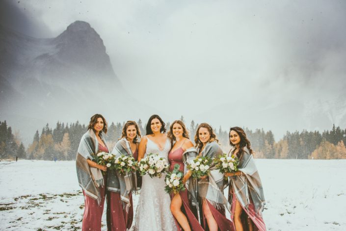 Meagan and her bridesmaids standing in a snowy field in the Rocky Mountains. The bridesmaids are wearing mauve dresses and blanket scarves. They are all carrying mauve, blush and white bouquets with greenery. Blush and Mauve Canmore Wedding.