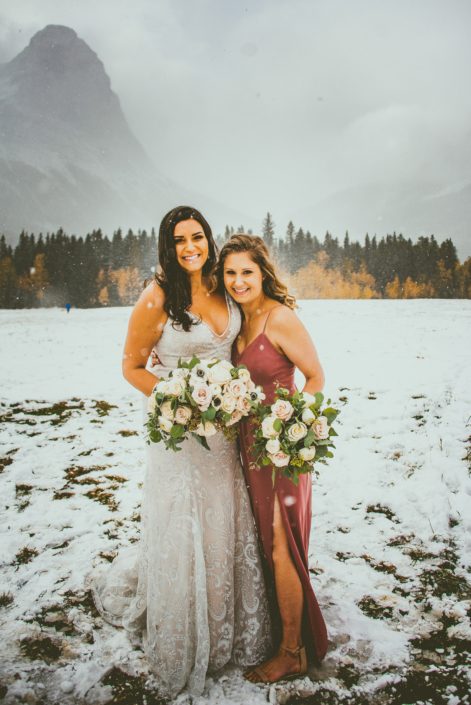 Bride and Bridesmaid standing in the snow carrying bouquets made of blush, white and mauve roses with greenery.