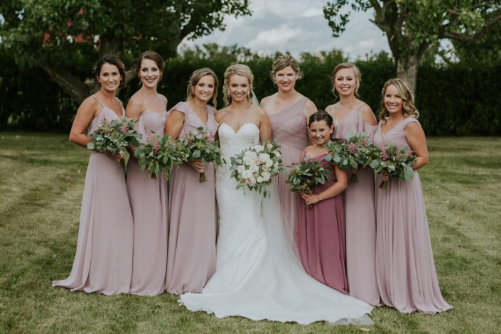 Rustic Chic Blush Wedding - Brooke wearing a white lace bridal gown and holding a blush and ivory bouquet made of roses, ranunculus and eucalyptus surrounded by her bridesmaids wearing blush floor length gowns and holding eucalyptus greenery bouquets with a touch of pink astilbe.