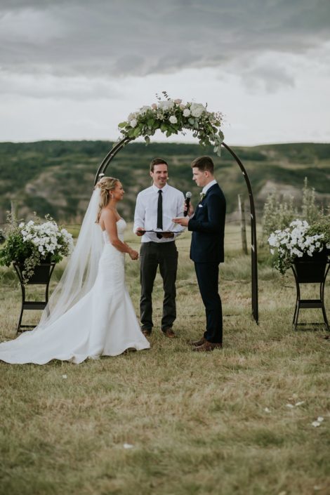 Bride and groom, Brooke and Levi, standing under an archway adorned with a flower arrangement made of white hydrangea, white o'hara garden roses, playa blanca roses, quicksand roses, salal, Italian ruscus and eucalyptus greenery.
