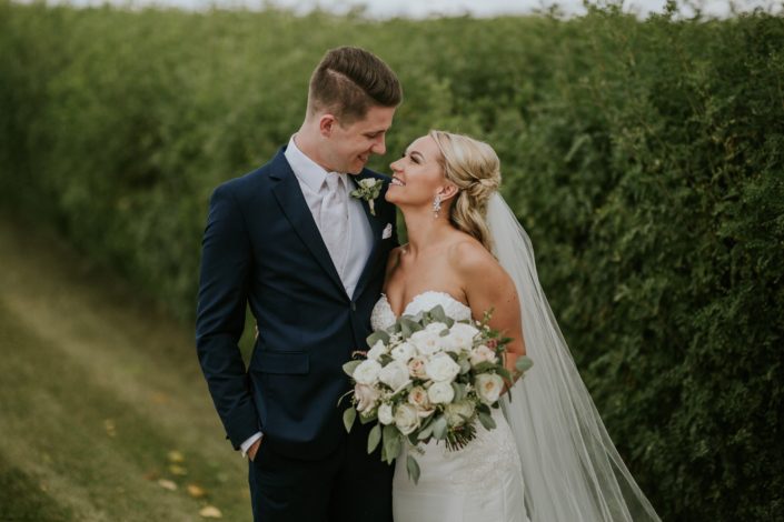 Brooke and Levi smiling at each other while holding a blush and ivory bouquet made of white o'hara garden roses, quicksand roses, playa blanca roses, astilbe, waxflower and eucalyptus greenery.