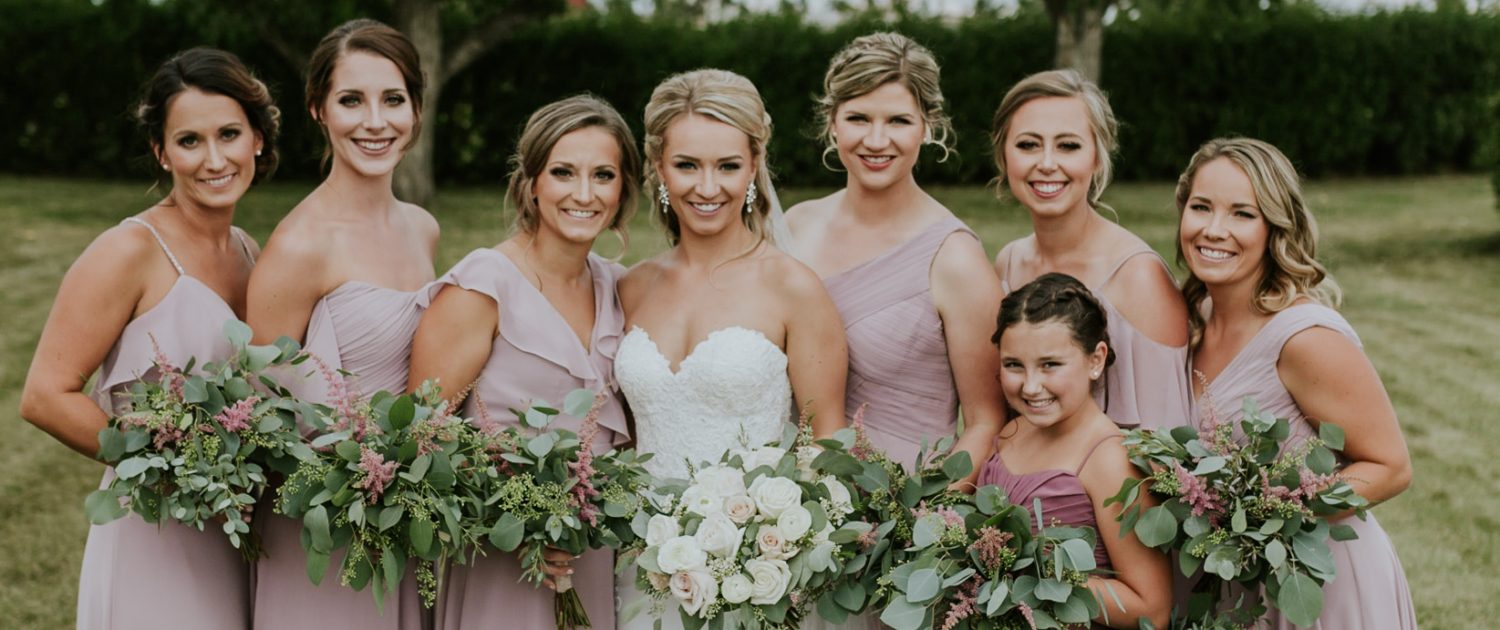 Bride, Brooke, wearing a white lacey dress and holding a blush and ivory bridal bouquet made of white o'hara garden roses, quicksand roses, playa blanca roses, ranunculus, astilbe, wax flower and eucalyptus. Standing with her bridesmaids and jr. bridesmaid wearing blush gowns and holding eucalyptus bouquets with a touch of pink astilbe.