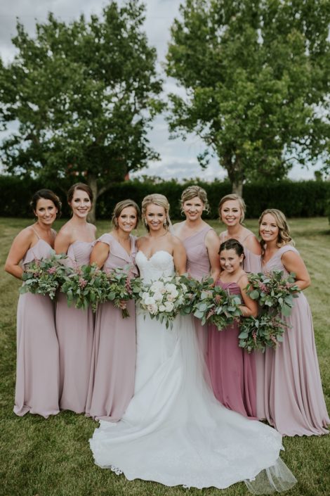 Bride, Brooke, wearing a white lacey dress and holding a blush and ivory bridal bouquet made of white o'hara garden roses, quicksand roses, playa blanca roses, ranunculus, astilbe, wax flower and eucalyptus. Standing with her bridesmaids and jr. bridesmaid wearing blush gowns and holding eucalyptus bouquets with a touch of pink astilbe.