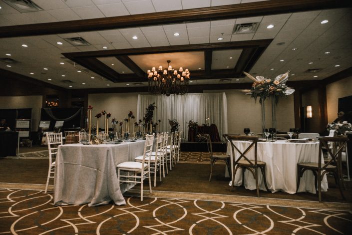 Cambridge Bridal Show 2020 - long table with white chairs decorated with tall gold centrepieces made of red roses and round tables with wood chairs and tall structural arrangement with metallic dyed Anahaw palm leaves, pampas grass, blush roses and greenery.