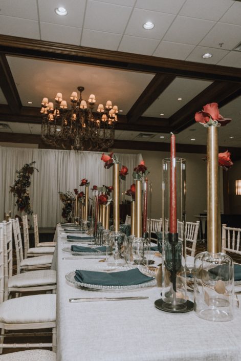 Cambridge Bridal Show 2020 - long table with white chairs, blue napkins and tall gold cylinder vases and red roses.