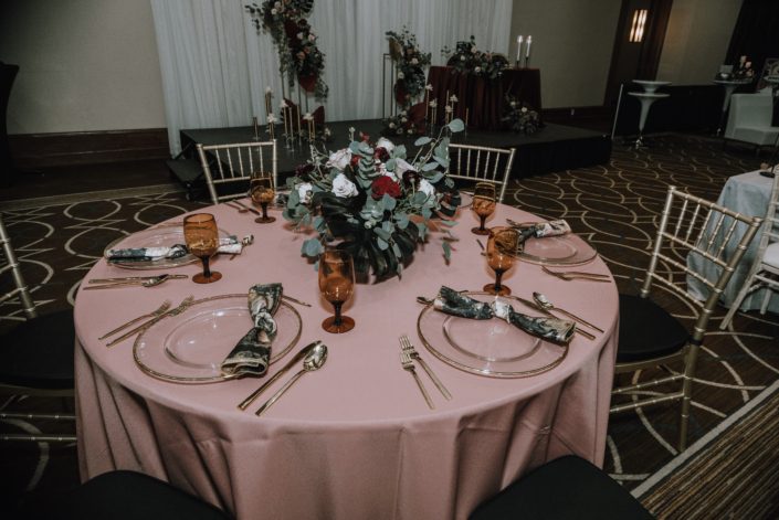 Cambridge Bridal Show 2020 - Round tablescape with blush pink table cloth and low centrepiece made of black dyed monstera leaves, red and blush roses, and eucalyptus greenery.