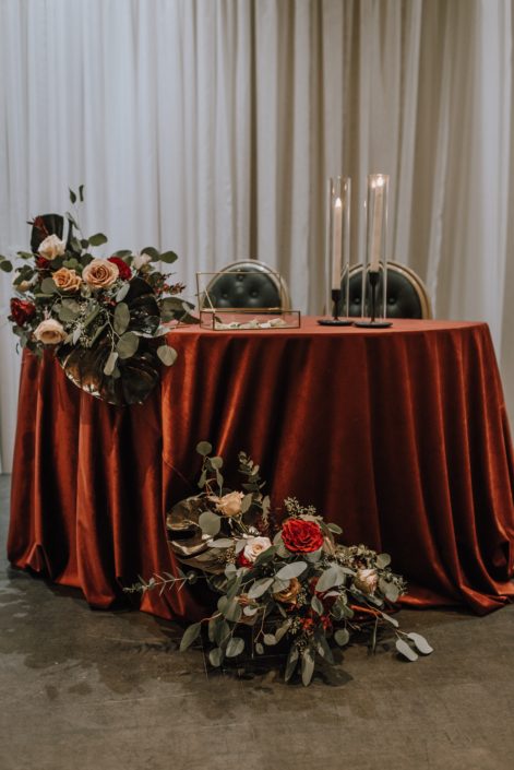 Cambridge Bridal Show 2020 - small couple table covered with red tablecloth decorated with arrangements made of dyed monstera leaves, red and blush roses and eucalyptus greenery.