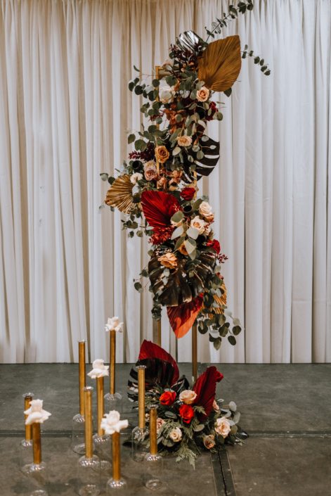 Cambridge Bridal Show 2020 - dramatic tall vertical arrangement made of black monstera leaves, red and metallic dyed Anahaw palm leaves, red and blush roses, and eucalyptus greenery.