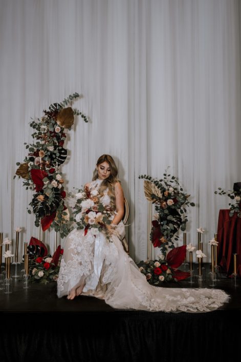 Cambridge Bridal Show 2020 - model wearing lace bridal gown holding bridal bouquet made of pampas grass, red and blush roses, ranunculus, and eucalyptus greenery tied with trailing ribbon. She is surrounded by dramatic tall vertical arrangements made of black monstera leaves, red and metallic dyed Anahaw palm leaves, red and blush roses and eucalyptus greenery.