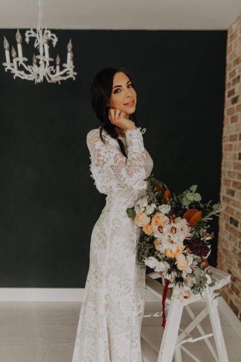 Neutrals Styled Shoot with Down the Aisle - Bride wearing lace gown standing on a ladder holding a neutral coloured bouquet with orchids, peach roses, magnolia leaves and eucalyptus, tied with trailing ribbons.
