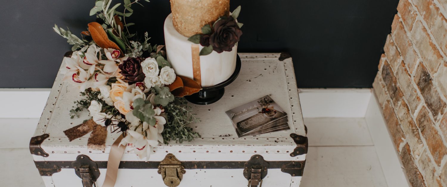 Neutrals Styled Shoot with Down the Aisle - Cake and neutral coloured bouquet with orchids, roses, magnolia leaves and eucalyptus greenery on top of a vintage trunk.