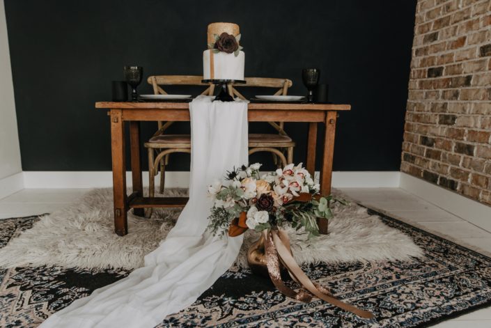 Neutrals Styled Shoot with Down the Aisle - Neutral coloured bridal bouquet with roses, orchids, magnolia leaves and eucalyptus greenery tied with trailing ribbons next to a decorated sweetheart table.