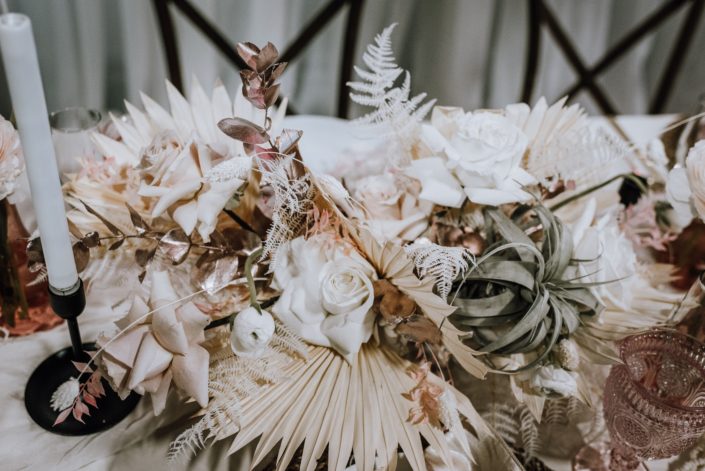 Close up of a centrepiece made of cream coloured dried foliage, white roses, metallic dyed eucalyptus, bunny tails and an airplant.