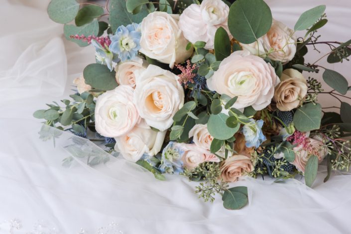 Up close shot of Amy's Pink and Blue bridal bouquet featuring white o'hara garden roses, quicksand roses, blush ranunculus, blue delphinium, eryngium, astilbe and a mixed variety of eucalyptus greenery.