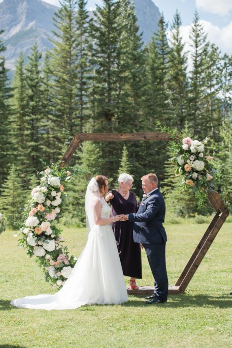 Amy and Kerry standing in front of the wooden hexagon archway at the ceremony. The archway is decorated with fresh floral arrangements designed with white hydrangeas, peach chrysanthemums, white o'hara garden roses, quicksand roses, astilbe, salal and eucalyptus.
