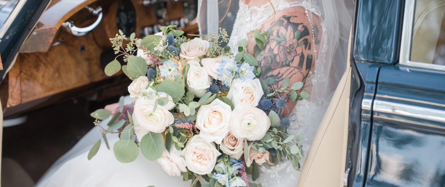 Amy in a vintage car holding her pink and blue bridal bouquet featuring white o'hara garden roses, quicksand roses, pale pink ranunculus, astilbe, blue delphinium, eryngium, and eucalyptus.