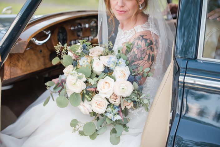 Amy in a vintage car holding her pink and blue bridal bouquet featuring white o'hara garden roses, quicksand roses, pale pink ranunculus, astilbe, blue delphinium, eryngium, and eucalyptus.