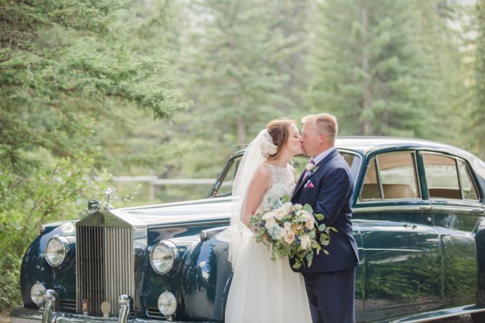 Amy and Kerry kissing in front of a vintage car. Amy is holding a pink and blue bridal bouquet made of white o'hara garden roses, quicksand roses, ranunculus, delphinium, astilbe, eryngium, and eucalyptus. Kerry is wearing a boutonniere made of white spray roses, blue delphinium, astilbe, eryngium and eucaluptus.