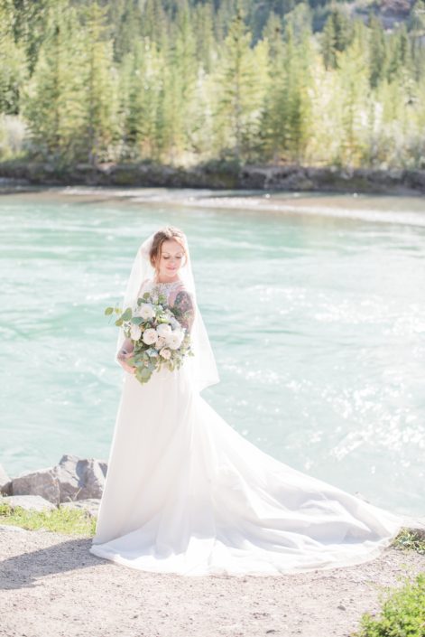 Amy standing on a river bank near Canmore in the Rocky Mountains while holding a pink and blue bridal bouquet featuring white o'hara garden roses, quicksand roses, ranunculus, delphinium, eryngium, astilbe, peach chrysanthemums, and eucalyptus.