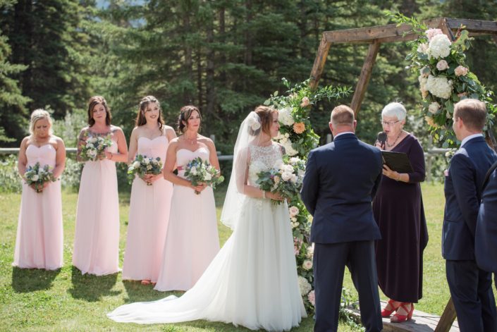 Bride and bridesmaids standing by the wooden hexagon archway at the wedding ceremony. They are all carrying bouquets made with white o'hara garden roses, quicksand roses, pale pink ranunculus, astilbe, eryngium, and a mixed variety of eucalyptus greenery.