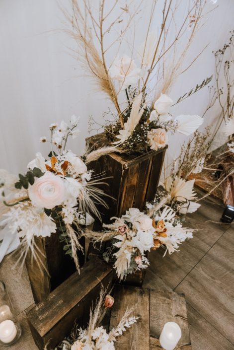 Arrangements on wooden crates at the Down the Aisle Wedding Show 2020 designed with dried leaves and branches, white playa blanca roses, orchids, pale pink ranunculus and pampas grass.