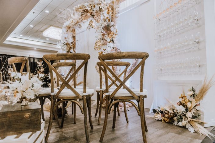 Wooden chairs and wall with champagne glasses and floral arrangement made of pampas grass, white playa blanca roses, orchids, gold painted monstera leaves, and other dried foraged leaves and branches.