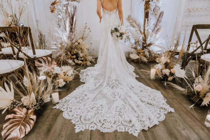 Lace bridal gown train surrounded by arrangements made of gold painted monstera leaves, painted sago palm, pampas grass, playa blanca roses, quicksand roses and other flowers and dried foraged leaves and branches.