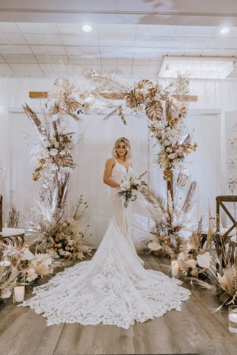 Model holding a white and blush bouquet made of roses and eucalyptus standing under a wooden arch and surrounded by arrangements made of gold painted monstera leaves, sago palm, pampas grass, and other flowers and dried leaves and branches.