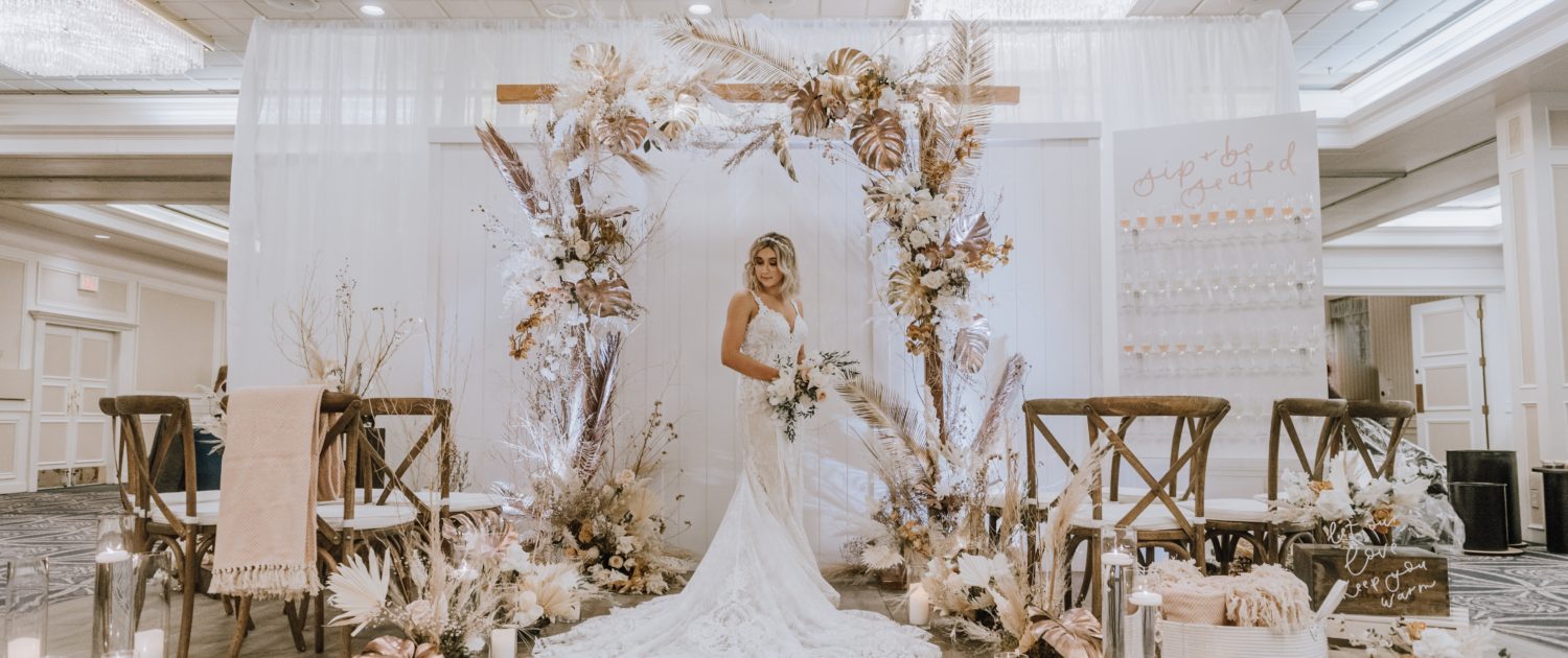 Down the Aisle Wedding Show booth decorated with gold painted monstera leaves, painted sago palm, pampas grass, white and pale pink roses, and other dried leaves and branches. Model wearing lace bridal gown and holding a blush and white bridal bouquet.