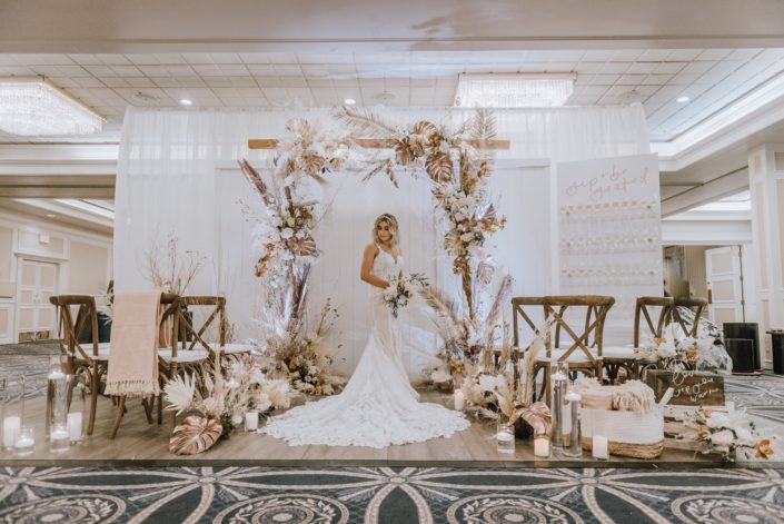 Down the Aisle Wedding Show booth decorated with gold painted monstera leaves, painted sago palm, pampas grass, white and pale pink roses, and other dried leaves and branches. Model wearing lace bridal gown and holding a blush and white bridal bouquet.