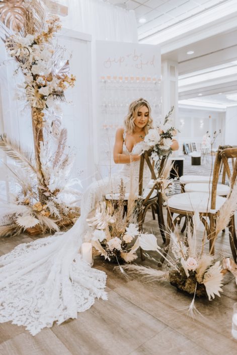 Model at the Down the Aisle Wedding Show holding a white and blush bouquet with roses, Japanese sweet peas and eucalyptus greenery surrounded by arrangements made of blush pink roses, gold painted monstera leaves, pampas grass and dried leaves and branches.