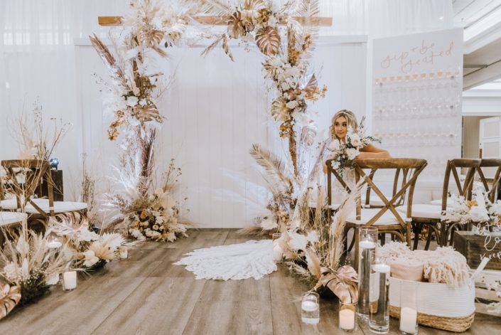 Down the Aisle Wedding Show 2020 model sitting and holding pale pink and white bouquet surrounded by arrangements made of dried leaves and branches, pampas grass, metallic gold painted monstera leaves, Japanese white sweet peas, quicksand roses, and painted sago palm.