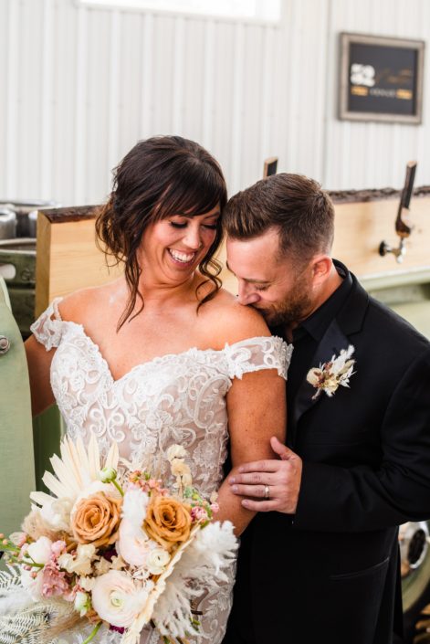 Groom kissing bride's shoulder at the Prohibition Pickup Photoshoot. Groom is wearing a boutonniere made of dried botanicals and the bride is holding a dusty pink coloured bouquet made with dried palm, pampas grass, ranunculus and roses.
