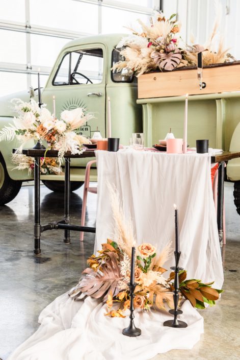 Sweetheart table draped with ivory linens and decorated with pillar candles, tableware, and floral arrangements designed with dried palm, pampas grass, metallic painted monstera leaves, roses and magnolia leaves.