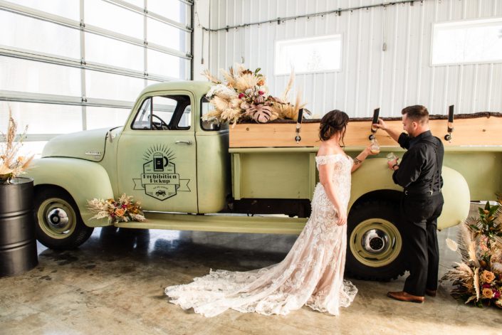 Bride and groom pouring beer from the Prohibition Pickup surrounded by floral arrangements made with dried palm, pampas grass, metallic painted monstera leaves and a variety of dusty pink coloured flowers.