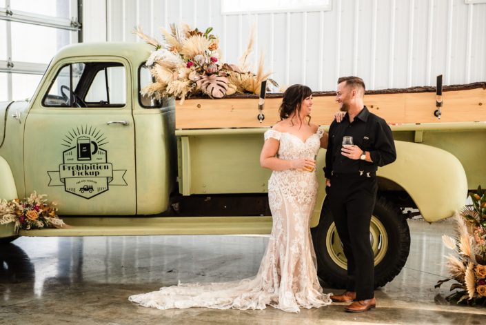 Bride and groom holding beer in front of the prohibition pickup that is decorated with a large floral arrangement made of dried palm, pampas grass, metallic painted monstera leaves and a variety of dusty pink flowers,