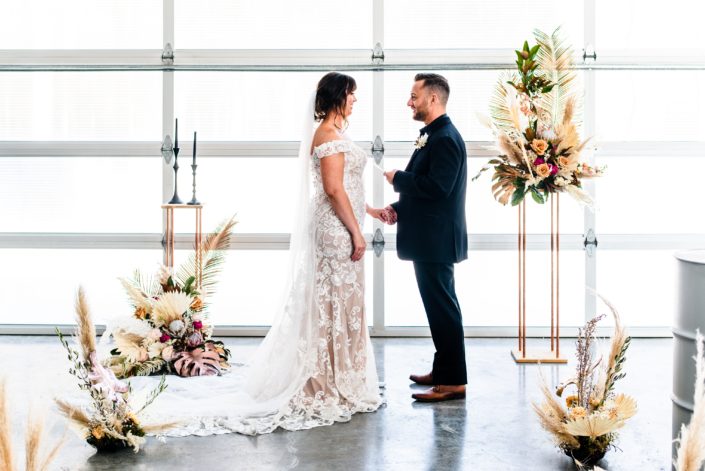 Bride and groom during the ceremony surrounded by floral arrangements with metallic painted monstera leaves, roses, pampas grass and other dried botanicals.