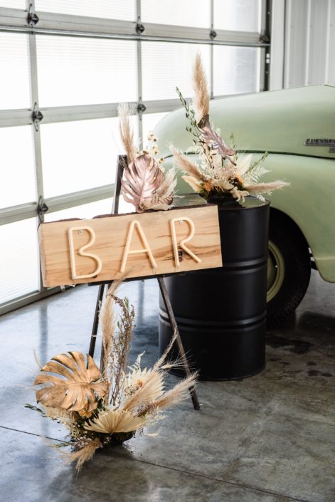Wooden "bar" sign surrounded by dried floral arrangements made of metallic painted monstera leaves, dried palm and pampas grass.