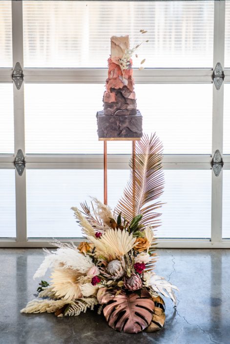 Cake from Sugar by Tracy and large floral arrangement made of metallic painted monstera leaves, dried palm, pampas grass, and dusty pink coloured florals.