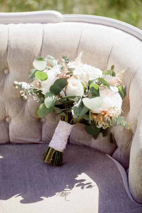 Cream and Blush Vintage Chic Bridal Bouquet on an antique linen tufted sofa; bouquet is made of white o'hara garden roses, quicksand roses, pale pink astilbe, babies breath, white peonies, dusty miller and eucalyptus wrapped with a blush satin and ivory lace overlay.