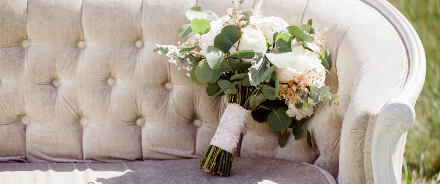 Cream and Blush Bridal bouquet propped on a linen tufted sofa; bouquet features white peonies, white o'hara garden roses, quicksand roses, astilbe, babies breath, dusty miller and eucalyptus with a blush satin and ivory lace overlay wrapped handle.